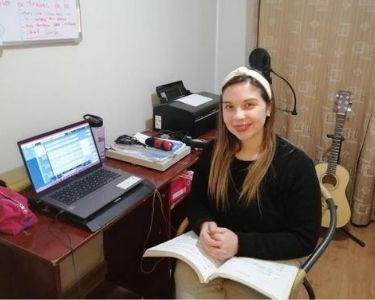 Una profesora está en su lugar de trabajo online, en donde destaca un notebook, una guitarra y sus partituras