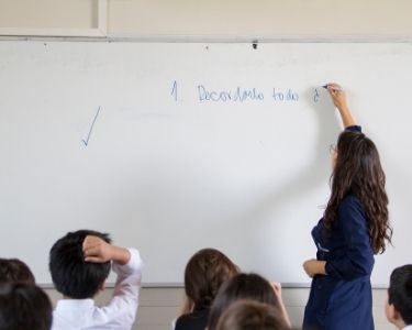 Imagen de una profesora de espalda, escribiendo en la pizarrra. En primer plano se ven la cabezas de varios niños, atentos a lo que escribe la docente.